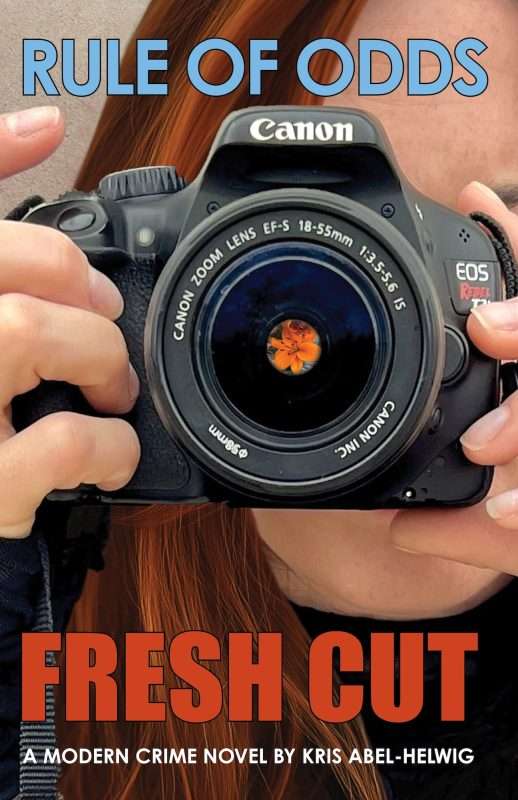 Book cover for a novel titled Rule of Odds: Fresh Cut. The design features a close-up photograph of a person holding a Canon EOS Rebel T3i camera directly toward the viewer. The camera dominates the center of the image. Reflected or composited inside the camera lens is a small orange flower, sharply contrasted against the dark lens. The person's hands, hair, and part of the face are partially visible behind the camera. At the top, large blue text reads “RULE OF ODDS.” At the bottom, large bold red text reads “FRESH CUT,” with smaller white text below stating “A Modern Crime Novel by Kris Abel-Helwig.” The overall tone is dramatic and photographic, suggesting a crime story linked to photography.